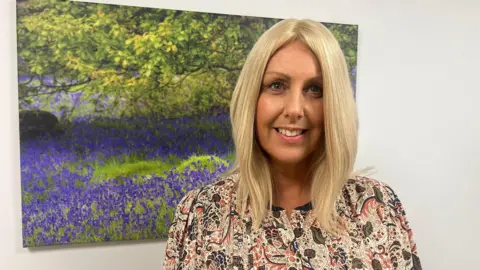 Manx Care chief executive Teresa Cope wears a brown top with a paisley design. She has shoulder-length blonde hair. She stands next to a large picture of bluebells.