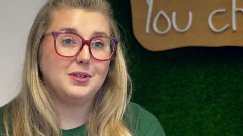 Lucy has long blonde hair and blue eyes. She is wearing redframed glasses and a green t-shirt.