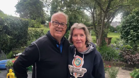 Paul is standing next to his wife.
He is wearing a blue top and glasses and standing in his back garden.
There are trees and general green foliage in the background.