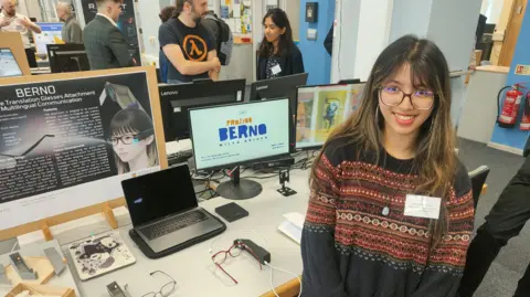 A woman wearing a knitted jumper and glasses sits in front of a computer screen while smiling at the camera. Another pair of glasses attached to a device can be seen on the table. 