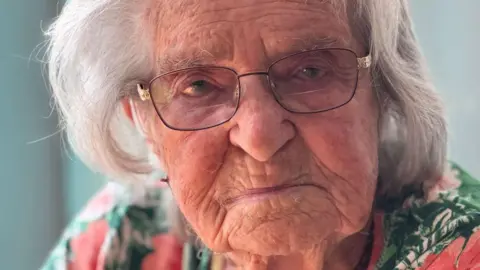 An elderly woman with grey hair, glasses and a floral patterned top