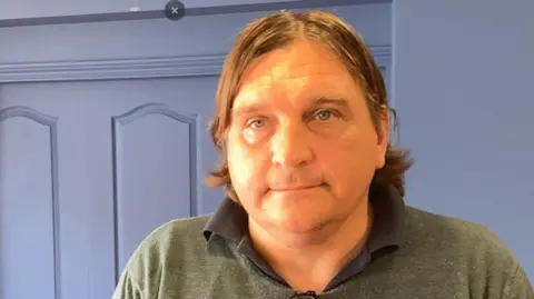 Stuart Austin. He has medium length brown hair and is wearing a dark green jumper over a black shirt.