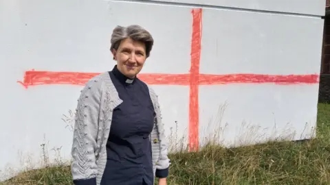 LDRS Reverend Rachel Heskin (woman with short hair, blue top and grey cardigan. There is a red cross painted on a white wall behind her). There is also long grass.