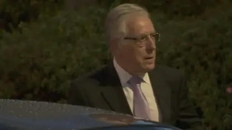 O'Brien has grey hair combed loosely back and wears glasses. He is wearing a dark suit jacket and a lilac tie. 