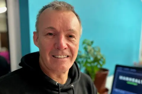 Paul has short greying hair and is wearing a black hoodie. He is pictured in front of his laptop and is smiling. 