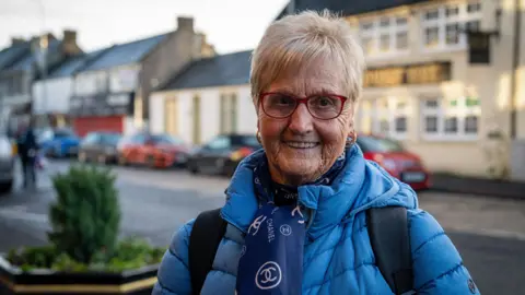Susan Snow, a retired lady with short blonde hair and glasses smiles at the camera in the local main street, wearing a blue winter jacket and a blue and white neck scarf. There is a pub behind her but the name sign is blurred.