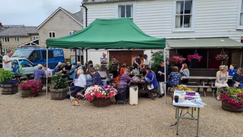 Sax Community Fridge A crowd of people sat on benches outside a pub with lots of planters full of flowers