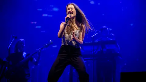 Sigrid onstage - she is singing into a microphone while her band play behind, bathed in blue light.