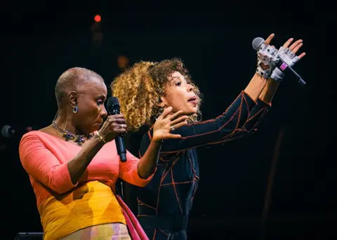 PAT BATARD / HANS LUCAS / AFP / GETTY IMAGES Two women clap their hands and dance on stage facing the audience.