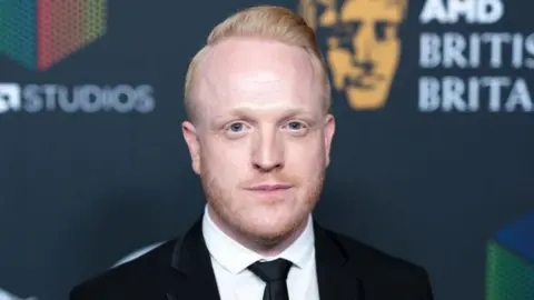  Owain Rhys Davies wearing a black blazer, black tie and white shirt.
