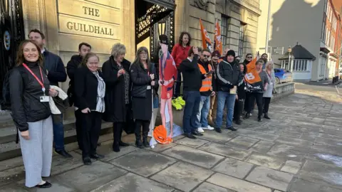 GMB Union Several people are standing next to the steps of The Guildhall in Hull. Some are holding orange and white GMB Union flags and there is a cardboard cutout of David Hasselhoff in the middle.