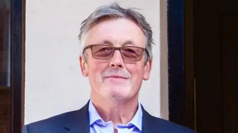 Shane John Gerard Connolly pictured with a neutral expression on his face. He is wearing rectangular shaped glasses, he has grey hair and is wearing a white collar shirt underneath a grey jacket.