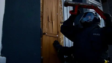 Merseyside Police A police officer in protective clothing and helmet about to hit a door in a raid