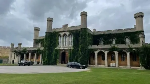 Exterior of Lincoln Crown Court. It is a two-storey Victorian Gothic building with crenellated towers and arches and foliage growing up the walls. In front is a grass lawn and a roadway with two cars parked on it.