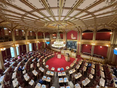 Interior of the Storting. It is looking down on dozens of desks arranged in semi-circular rows in an ornate room with red carpets and gold panelling
