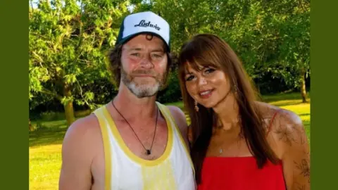 Lee Strange Take That's Howard Donald on the left in a white and yellow tank top and cap and Camilla is on the right dressed in red. They stand in front of trees