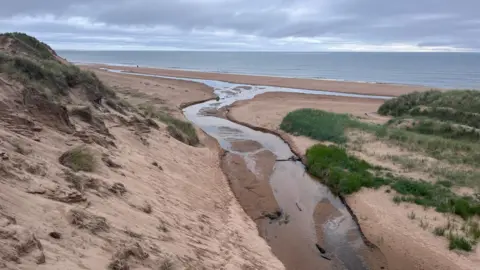 A view of a sandy beach with the sea beyond and a small creek of water running through the middle of the shot to the sea.