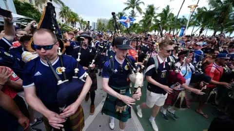 Thousands of Scotland fans marching along a boulevard, with several playing bagpipes. Nearly all of the fans are wearing Scotland football shirts. 