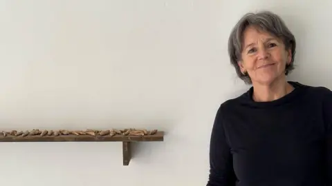 Helen Acklam Helen stood to the right, against a white wall. She is smiling. There is a shelf to her left with pieces of her artwork displayed.