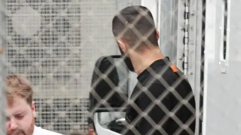 Eddie Mitchell A man in a white uniform is next to a man in dark clothing. They are pictured behind a grey wire fence.