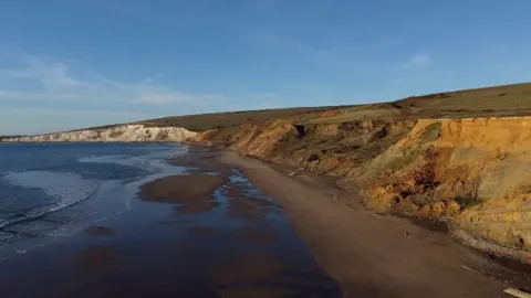 Late evening sunshine gives the crumbling cliffs of Compton beach and orange glow with deep blue waters and white chalk cliffs in the background