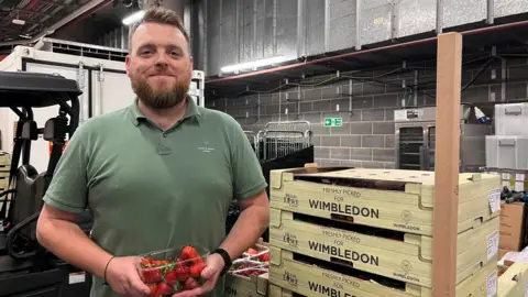 Ryan Stanton in a green polo shirt holding a punnet of strawberries. He is stood next to stacked up trays, labelled "freshly picked for Wimbledon", in a warehouse.