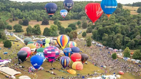 A picture of many hot air ballons on a large field. Some are in the air and others are on the ground. There are large crowds of poeple