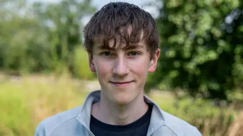 Harris McCutcheon, teenager looking at camera, wearing a light grey top over a black t-shirt, with grass and trees in the background