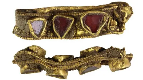 The British Museum Two views of a gold and garnet early medieval ring, still encrusted with mud after coming out of the earth. The ring has been flattened over the centuries. It has seven raised red semi-precious stones set into the band. 