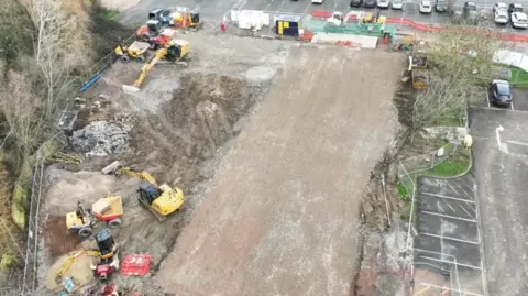 NHS North West Anglia NHS Foundation Trust An aerial shot of a building site. In the middle is a flattened area and on the left there is a yellow front loader. Two yellow excavators are levelling the site. At the top a car park with cars can just be seen. 