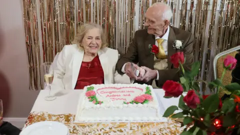 Andrea and Jimmy sat at a table, preparing to cut their wedding cake. Jimmy is smiling at his new bride and she is smiling at the camera.
