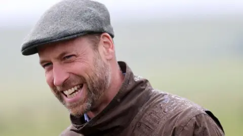 Prince William wearing a brown wax coat and gray wool cap. He has a beard and is smiling at the camera