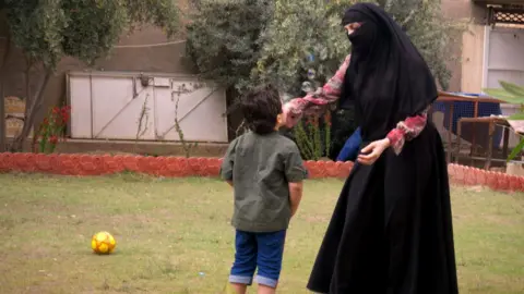 Safa plays with her young son in the garden. He is wearing a green shirt and short jeans and is blowing bubbles. She is wearing a long black skirt, a red patterned top with long sleeves and a black headscarf and veil covering most of her face. There is a yellow ball on the grass nearby.
