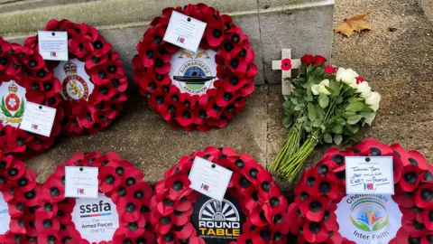 Several poppy wreaths, a bouquet of flowers and a crucifix cross on the ground. The wreaths have notes attached to them and the logo of the organisation they came from.