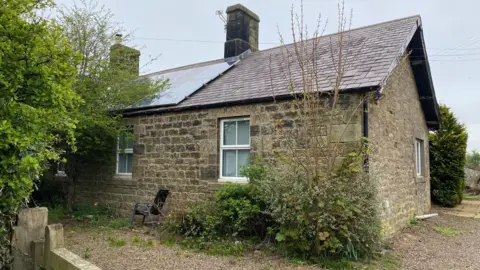 East Cottage is a traditional single-storey stone built cottage and about 200 years old. There are well established bushes near it and a double row of solar panels on the pitched roof.