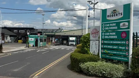 BBC/Chris Long The entry to Roberts Bakery, where a green sign with directions to the car park and deliveries stands in front of a gate and a green security hut. In the background, a green and dark brown shed stands with a white Roberts-liveried lorry moving out of it and other factory buildings can be seen