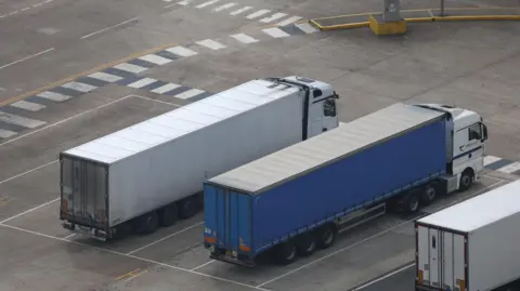 Three lorries, parked side-by-side a vehicle width apart in desginated spaces. Two are white and the middle one is blue.