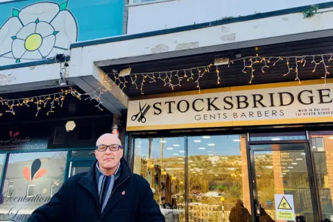 A man with a bald head and glasses is wearing a scarf and a smart winter coat. He is standing in front of a barber's shop with a sign saying Stocksbridge Gents Barbers. There is also a large white Yorkshire Rose on a blue banner behind him