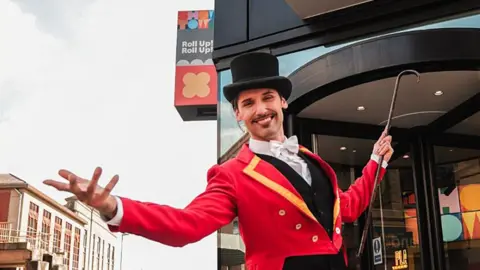 A man with a moustache in a ringmaster's costume - including a top hat, red jacket and cane - poses with outstretched arms and a big smile outside the museum
