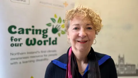 A woman with short curly blonde hair smiles at the camera. She has a black top on, earrings and red lipstick 