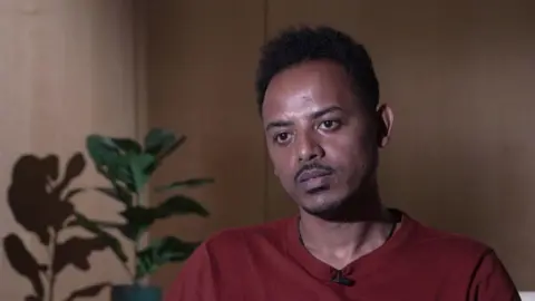 A man with black moustache and hair wearing a maroon t-shirt. He is sitting in a room with wooden cladding walls and a plant in the background.