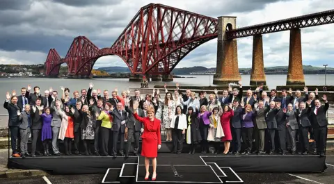 Getty Images Nicola Sturgeon, in a red blazer and dress, stands with the 56 SNP MPs who won seats in the 2015 election. They are in front of the Forth Road Bridge.