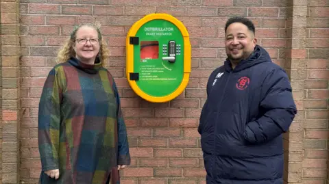 A woman with a multicoloured gown and curly blonde hair stands beside a man with short dark hair wearing a waterproof sports coat. Behind them is a green and yellow plastic case with a red defibrillator inside.
