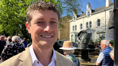 Matthew Ward, smiling and wearing a shirt and blazer, with people and a fountain behind him.