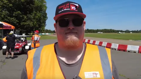 Drew Hanslow has a cap, sunglasses and an orange hi-vis on. Behind him is a race track.