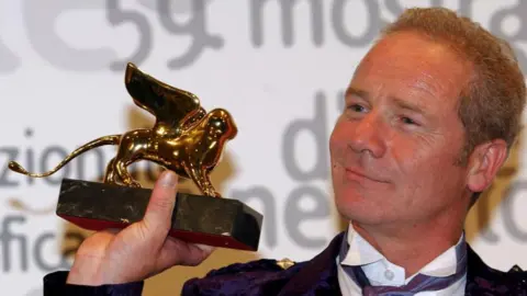 Getty Images Peter Mullan, clean shaven this time, holds the Golden Lion trophy up and looks at it. He is wearing a dark purple kilt jacket with white shirt and mauve cravat.