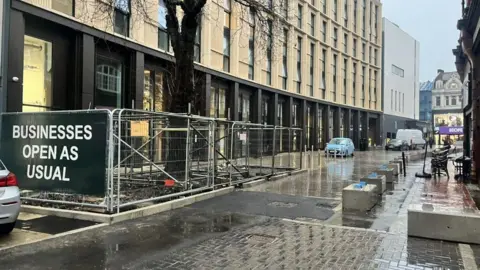 Rachel Russell/BBC A general view of Central Road with a sign saying "businesses open as usual" on some metal fencing that is covering construction work. The street appears mostly empty due to rain. 