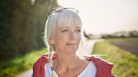 A woman goes on a walk in the countryside. She has her hair in plaits and is wearing a white T-shirt with a red layer. Grass and hedges and a road can be seen behind her in the close-up picture. She has glasses on the top of her head.