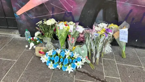 LDRS A number of bunches of flowers on a pavement next to a wall which has been painted with a bright, multi-coloured scene of people dancing.