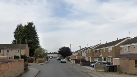 A road running between two rows of houses. Cars are parked on the street. The homes are mainly semi-detached with fences separating homes and the odd car parked on a driveway.